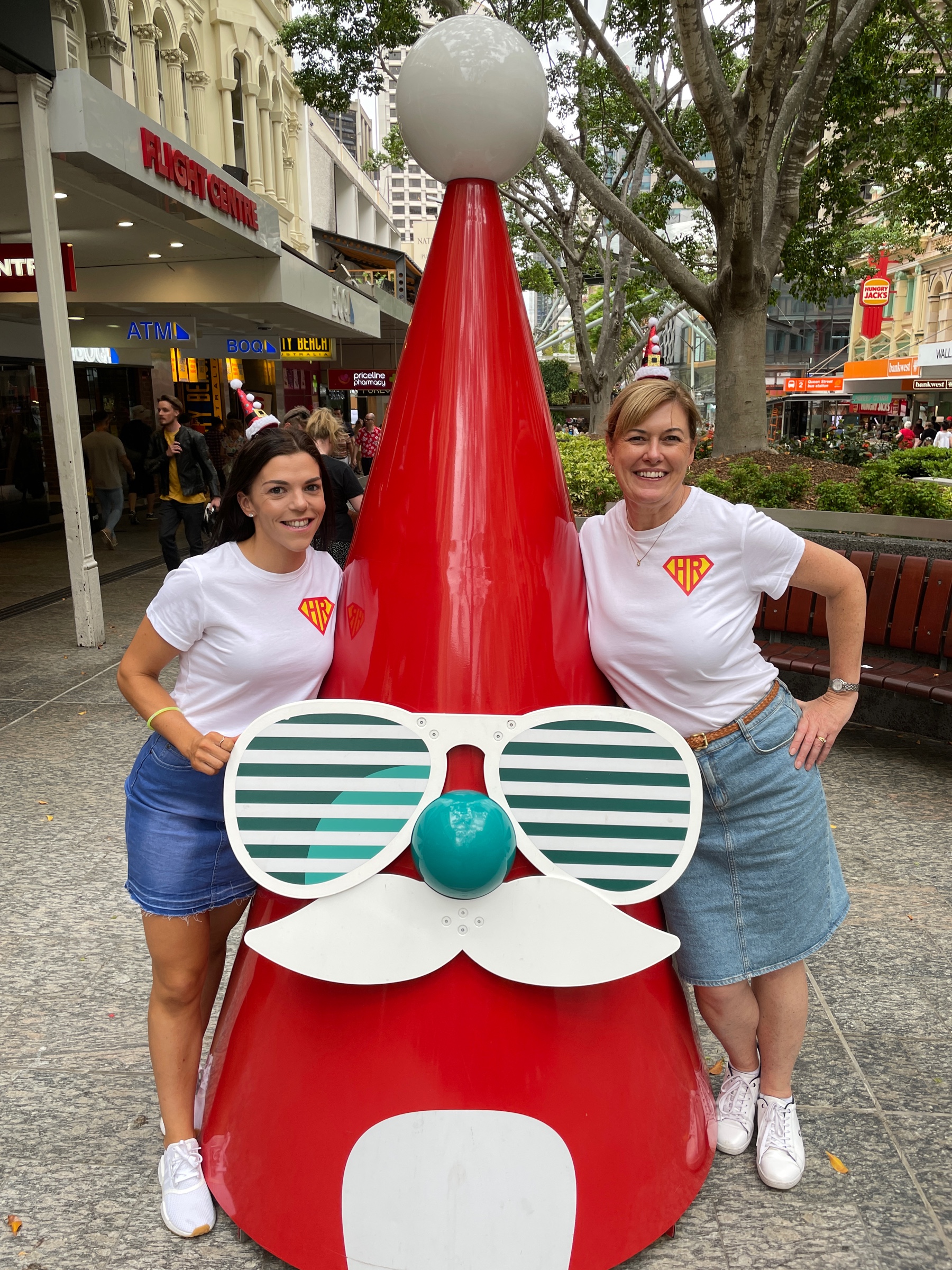 Two women smiling beside a giant red Santa sculpture during a Christmas scavenger hunt on the Gold Coast.